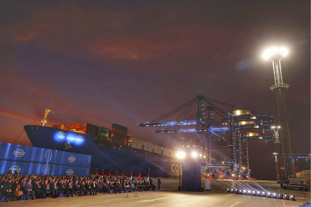 A cargo ship branded Cosco Shipping is docked at the Chancay megaport. A crowd sits waiting for a speech.